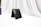 A black leather wallet is positioned upright on a white surface with draped white fabric in the background. The wallet has a subtle embossed logo, and there is a blurred purple object to the left side.
