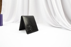 A black leather wallet is positioned upright on a white surface with draped white fabric in the background. The wallet has a subtle embossed logo, and there is a blurred purple object to the left side.