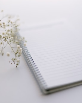 A serene notebook open beside a delicate sprig of jasmine on a soft off-white surface, with Arabic calligraphy softly blurred in the background.