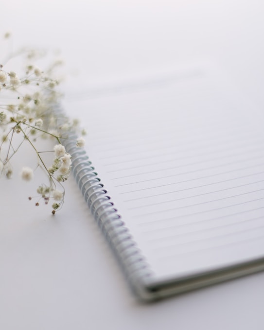 A spiral-bound notebook with lined pages is placed on a smooth surface, accompanied by delicate white flowers on the left side. The scene is softly lit, creating a calm and serene atmosphere.