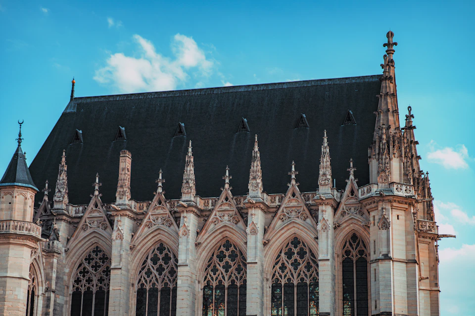 The image shows a large, Gothic-style church with a pointed roof and multiple spires reaching towards the sky. The church is constructed from stone and features numerous arched windows that add to its architectural charm. The perspective of the photo is taken from below, looking up at the imposing structure. The sky above the church is clear blue, providing a beautiful backdrop for this historic landmark.