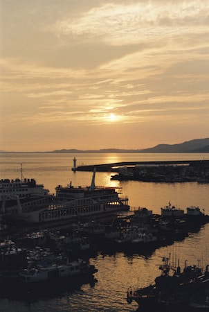 A peaceful harbor scene at sunset with boats docked and a lighthouse in the distance. The sun's warm glow reflects off the water, creating a tranquil and serene atmosphere.