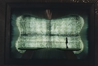 A silhouette of a person stands on a bridge-like structure inside a futuristic space with walls covered in glowing, grid-like patterns of light, creating an ethereal and immersive environment.