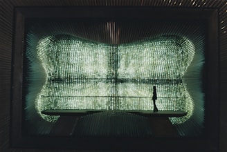 A silhouette of a person stands on a bridge-like structure inside a futuristic space with walls covered in glowing, grid-like patterns of light, creating an ethereal and immersive environment.