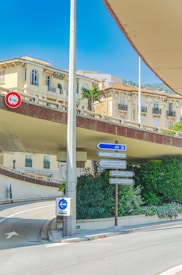 A street intersection with multiple road signs directs towards destinations like Monte Carlo and Cap d'Ail. The background features elegant, ornate buildings with arched windows, and there's a palm tree visible. The clear sky and bright sunlight create sharp shadows on the road.