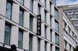 A modern building with multiple large glass windows arranged in a grid pattern. A vertical sign with the word 'HOTEL' is attached to the facade. The architecture is clean and minimalistic, with a focus on straight lines and uniformity.