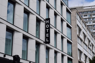 A modern building with multiple large glass windows arranged in a grid pattern. A vertical sign with the word 'HOTEL' is attached to the facade. The architecture is clean and minimalistic, with a focus on straight lines and uniformity.