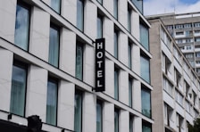 A modern building with multiple large glass windows arranged in a grid pattern. A vertical sign with the word 'HOTEL' is attached to the facade. The architecture is clean and minimalistic, with a focus on straight lines and uniformity.