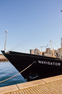 A large black boat with the name 'Navigator' is moored by a cobblestone dock. The clear blue sky stretches above, and a seagull perches on a wooden pole extending from the boat. Modern architectural buildings and cranes can be seen in the distant background across the water.