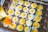 A batch of small sunny-side-up eggs is cooking on a griddle with circular molds. Some eggs are in the process of being cooked and have slightly runny yolks. An orange-tipped bottle is squeezing oil or a similar liquid onto one of the eggs.