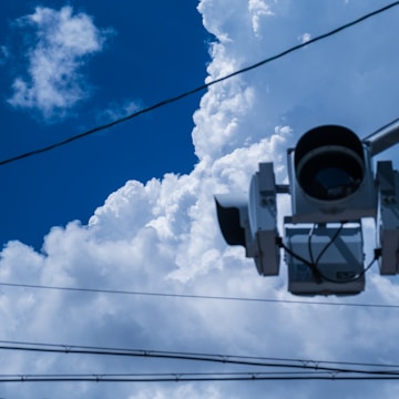 A traffic camera is observed against a backdrop of large, fluffy white clouds in a bright blue sky. Several utility wires run horizontally across the image.