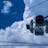 A traffic camera is observed against a backdrop of large, fluffy white clouds in a bright blue sky. Several utility wires run horizontally across the image.