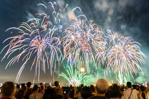 A large crowd gathers at night to watch a spectacular fireworks display. The sky is filled with bursts of colorful fireworks in shades of blue, green, red, and white, creating a vibrant and festive atmosphere.