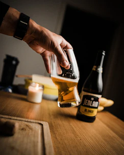 Close-up of hands holding freshly brewed craft beer glasses, with brewing equipment blurred in the background.