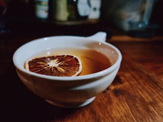 A warm beverage served in a white cup with a brown, dehydrated citrus slice floating on top. The cup sits on a wooden surface surrounded by a blurry background, possibly of kitchen items.