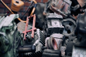 A collection of old, rusty mechanical parts and machinery pieces, piled together in a somewhat disorganized manner. The components include gears, bolts, a piston rod, and other indiscernible engine parts. The texture of the surfaces shows signs of wear and corrosion, with a layer of dust settled on top.