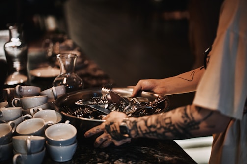 A tattooed person is handling a tray filled with silverware near a stack of white cups and a glass carafe, set against a dimly lit background with a rustic ambiance.