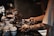 A tattooed person is handling a tray filled with silverware near a stack of white cups and a glass carafe, set against a dimly lit background with a rustic ambiance.
