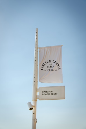 A tall white pole with a security camera attached supports a white flag and a rectangular sign. The flag displays the text 'Carlton Cannes Beach Club 1930' in a circular pattern. The rectangular sign below reads 'Carlton Beach Club.' The background features a clear blue sky.
