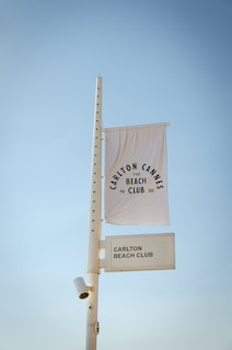 A tall white pole with a security camera attached supports a white flag and a rectangular sign. The flag displays the text 'Carlton Cannes Beach Club 1930' in a circular pattern. The rectangular sign below reads 'Carlton Beach Club.' The background features a clear blue sky.