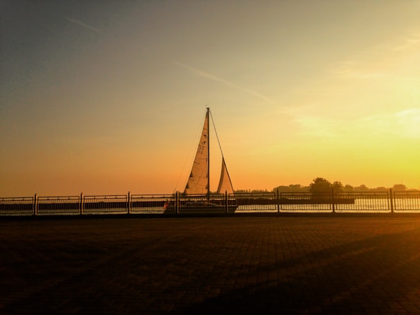 A sailboat moves smoothly on a calm body of water during sunset, bordered by a railing. The sky is filled with warm hues of orange and yellow, creating a peaceful and serene atmosphere.