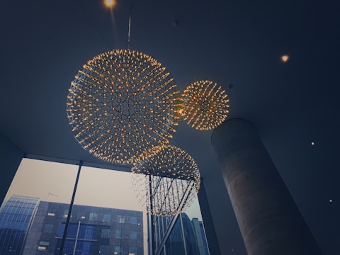 Three large spherical chandeliers with intricate designs are suspended from the ceiling in a modern interior space. The chandeliers emit a warm glow, contrasting with the dimly lit surroundings. Large windows in the background reveal a view of urban architecture.