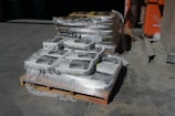 A stack of pallets loaded with bags of all-natural play sand, wrapped in plastic. The bags are labeled 50 pounds. A second pallet in the background contains similar packaging, possibly for different sand or concrete mix. The setting is outdoors, likely a construction or supply yard, with an orange equipment or storage container visible to the right.