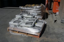 A stack of pallets loaded with bags of all-natural play sand, wrapped in plastic. The bags are labeled 50 pounds. A second pallet in the background contains similar packaging, possibly for different sand or concrete mix. The setting is outdoors, likely a construction or supply yard, with an orange equipment or storage container visible to the right.