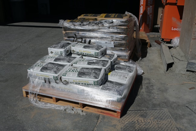A stack of pallets loaded with bags of all-natural play sand, wrapped in plastic. The bags are labeled 50 pounds. A second pallet in the background contains similar packaging, possibly for different sand or concrete mix. The setting is outdoors, likely a construction or supply yard, with an orange equipment or storage container visible to the right.