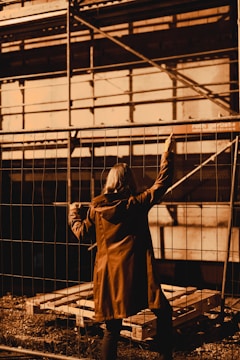 A person wearing a long coat stands facing a metal fence, with one arm reaching upwards. The scene appears to be a construction site, with scaffolding in the background and wooden pallets on the ground.