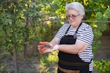 An older woman with short gray hair is standing outdoors in a garden, wearing a striped shirt and a black apron. She is holding soil in her gloved hands and is surrounded by green foliage and trees with small red berries.