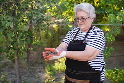 An older woman with short gray hair is standing outdoors in a garden, wearing a striped shirt and a black apron. She is holding soil in her gloved hands and is surrounded by green foliage and trees with small red berries.