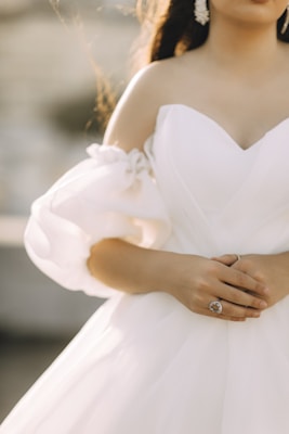 A close-up of a person wearing a white, off-shoulder wedding dress with intricate floral details on the long sleeves. The person is holding their hands together, showing a prominent, elegant ring. The background is softly blurred, providing a gentle contrast to the detailed fabric of the dress.