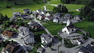 An aerial view of a small village nestled in a lush green landscape. The village consists of numerous houses with varied architectural designs, surrounded by well-maintained roads. A prominent church with a tall spire is situated in the center, adding a traditional element to the scene. The area is bordered by expansive fields and dense forest areas, providing a serene and picturesque environment.