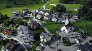 An aerial view of a small village nestled in a lush green landscape. The village consists of numerous houses with varied architectural designs, surrounded by well-maintained roads. A prominent church with a tall spire is situated in the center, adding a traditional element to the scene. The area is bordered by expansive fields and dense forest areas, providing a serene and picturesque environment.