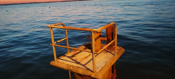 A rusted metal platform or structure is positioned over calm water, with a distant horizon where the water meets the sky. The platform appears to have a grated surface and handrails, suggesting it might be used for access or observation.