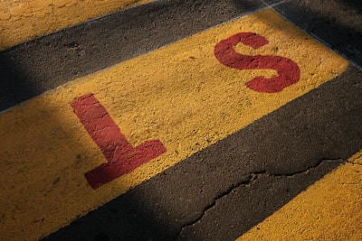 Yellow pavement with black stripes and large red letters 'L' and 'S' painted on it. The surface shows signs of wear and cracking.