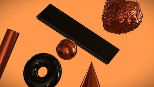 A collection of geometric shapes floats against an orange background. The objects include a shiny cylindrical shape, a black doughnut form, a metallic sphere, a smooth cone, a large rectangular block, and a complex, crumpled-texture sphere. Each shape has a reflective surface, giving a high-contrast, polished metallic appearance.