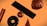 A collection of geometric shapes floats against an orange background. The objects include a shiny cylindrical shape, a black doughnut form, a metallic sphere, a smooth cone, a large rectangular block, and a complex, crumpled-texture sphere. Each shape has a reflective surface, giving a high-contrast, polished metallic appearance.