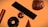 A collection of geometric shapes floats against an orange background. The objects include a shiny cylindrical shape, a black doughnut form, a metallic sphere, a smooth cone, a large rectangular block, and a complex, crumpled-texture sphere. Each shape has a reflective surface, giving a high-contrast, polished metallic appearance.