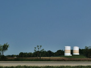 Two large, cylindrical storage tanks with horizontal orange bands are situated in a rural setting, surrounded by fields and trees. A road runs along the foreground, and the sky is clear and blue.