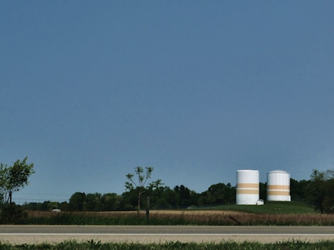 Two large, cylindrical storage tanks with horizontal orange bands are situated in a rural setting, surrounded by fields and trees. A road runs along the foreground, and the sky is clear and blue.