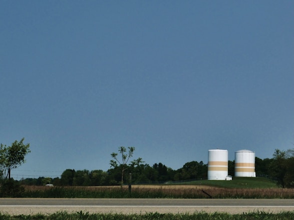 Two large, cylindrical storage tanks with horizontal orange bands are situated in a rural setting, surrounded by fields and trees. A road runs along the foreground, and the sky is clear and blue.