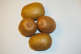 Four whole kiwifruit with brown, fuzzy skins are arranged close to each other on a light surface.