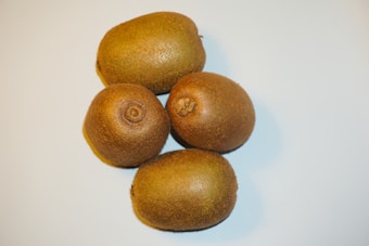 Four whole kiwifruit with brown, fuzzy skins are arranged close to each other on a light surface.
