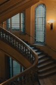 A beautifully designed spiral staircase with intricate metalwork on the railings, complemented by elegant stained glass windows with blue accents. The environment is softly lit with warm-toned light fixtures on the wall, contributing to a serene and classic atmosphere.