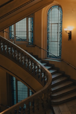 A beautifully designed spiral staircase with intricate metalwork on the railings, complemented by elegant stained glass windows with blue accents. The environment is softly lit with warm-toned light fixtures on the wall, contributing to a serene and classic atmosphere.