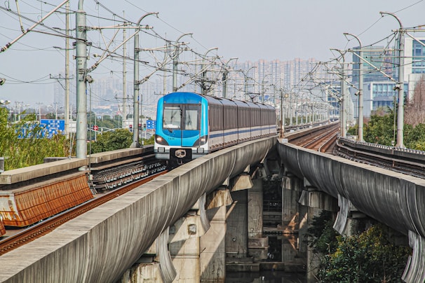 A blue and silver metro train travels on an elevated railway track with multiple power lines and poles parallel to it. The train is surrounded by urban city infrastructure, including buildings and greenery below the tracks. In the background, the skyline of high-rise buildings is visible.