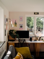 A cozy and neatly organized home office setup with a large window providing natural light. The desk features a computer monitor, a stylish chair, a desk lamp, a potted plant, and various decorative items. The wall above displays several colorful art prints. A small sofa with cushions sits near the desk, contributing to a comfortable and inviting atmosphere.