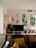 A cozy and neatly organized home office setup with a large window providing natural light. The desk features a computer monitor, a stylish chair, a desk lamp, a potted plant, and various decorative items. The wall above displays several colorful art prints. A small sofa with cushions sits near the desk, contributing to a comfortable and inviting atmosphere.